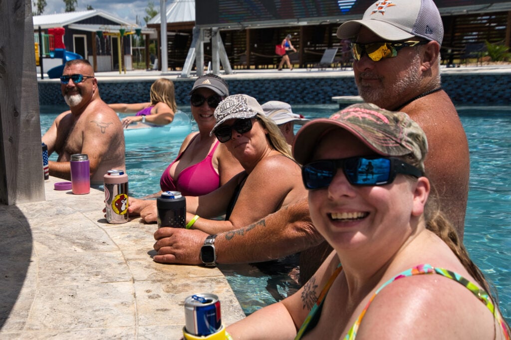 Five adults in swimsuits sit at the edge of a pool with drinks, smiling at the camera on a sunny day.