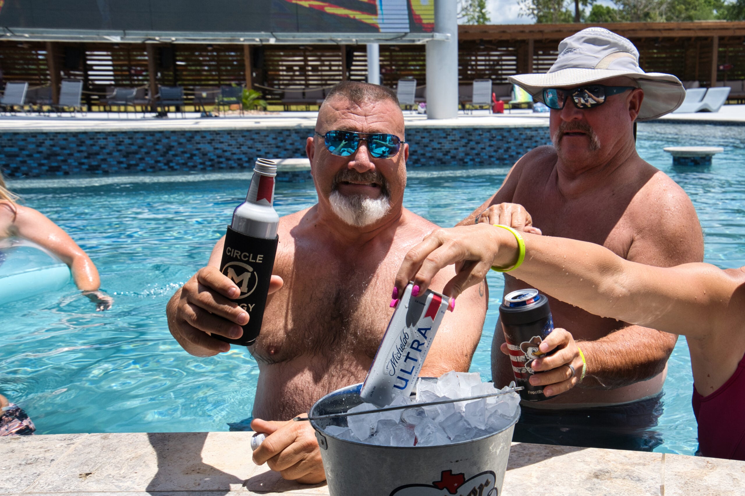 Two men in a pool hold drinks while a woman reaches for a can from a metal bucket filled with ice; all appear to be enjoying a sunny day.