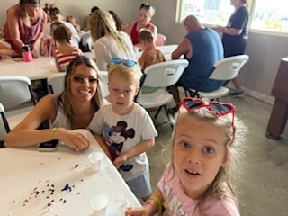 A woman and two children sit at a table with craft supplies, while other people work on projects at tables in the background.