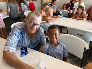 An older woman and a young boy sit together at a table in a busy room with other children and adults. The woman is smiling and holding a smartphone.