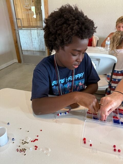 A child sits at a table sorting and stringing colorful beads from a container, with scattered beads on the white tabletop.