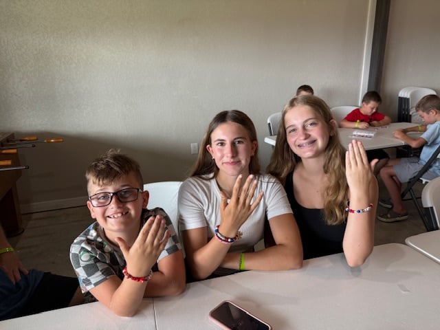 Three kids sit at a table, smiling and showing off handmade beaded bracelets. Other children are seated at tables in the background.