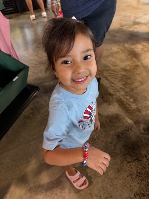 Young child with dark hair smiles at the camera, wearing a light blue shirt with a design, pink sandals, and a red, white, and blue beaded bracelet.