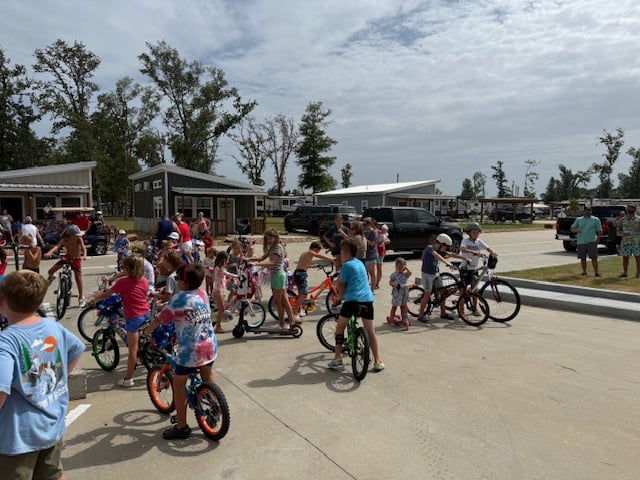 A group of children on bicycles gather on a paved street in a residential area with adults and small homes in the background.