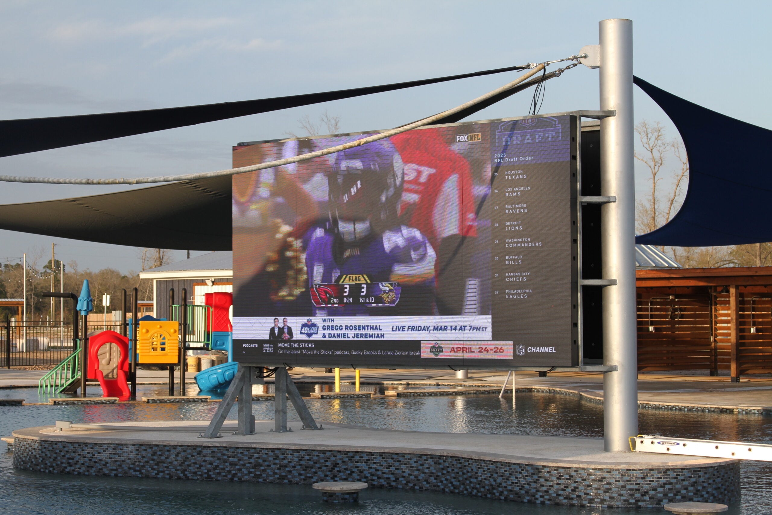 A large outdoor screen displays a football game above a pool area with playground equipment and pool-edge seating visible in the background.