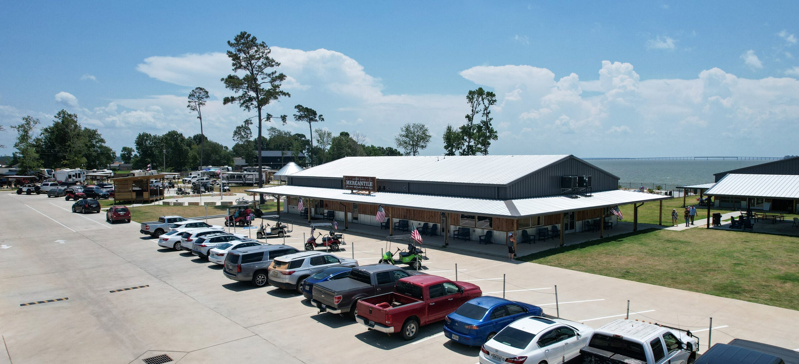 A row of vehicles is parked in front of a large, metal-roofed building labeled "Sharky’s Waterfront Grill," with trees, a grassy area, and water in the background.