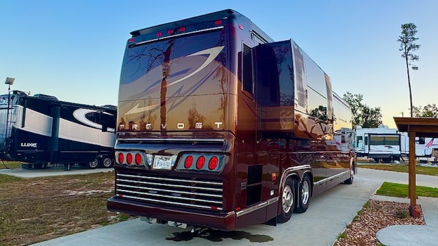 A large brown Prevost motorhome is parked on a paved site in an RV park, with other RVs and trees in the background.