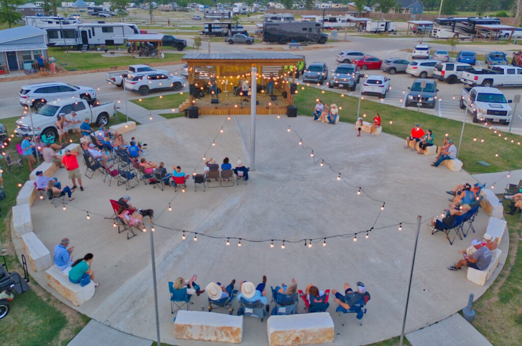 A group of people sit in a circular outdoor area, watching a band perform on a small stage, with string lights overhead and parked vehicles in the background.