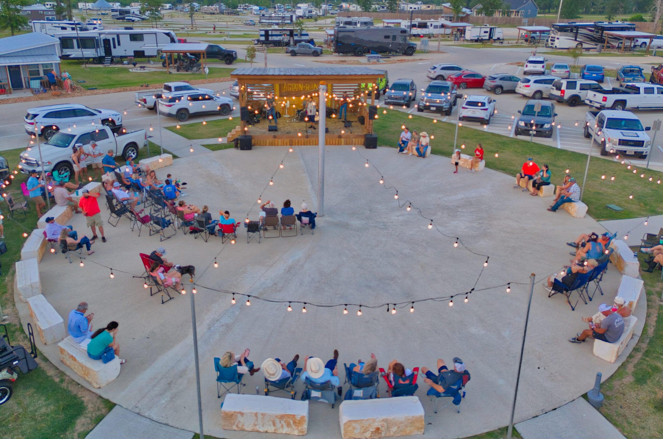 A group of people sit in a circular outdoor area, watching a band perform on a small stage, with string lights overhead and parked vehicles in the background.