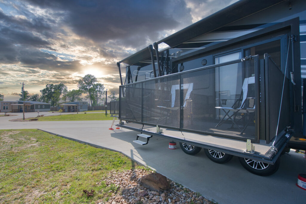 A modern RV with an outdoor seating area and awning is parked on concrete at sunset, with grassy surroundings and small buildings in the background.