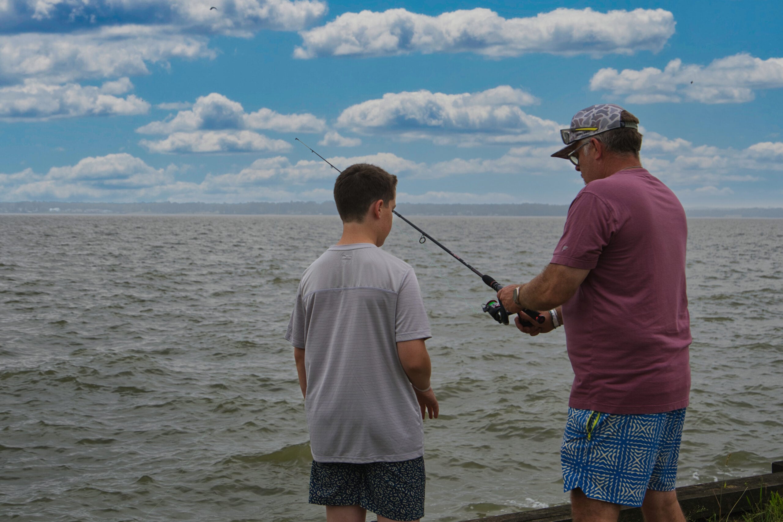 An adult and a child stand by the water's edge, with the adult holding a fishing rod and showing it to the child under a partly cloudy sky.