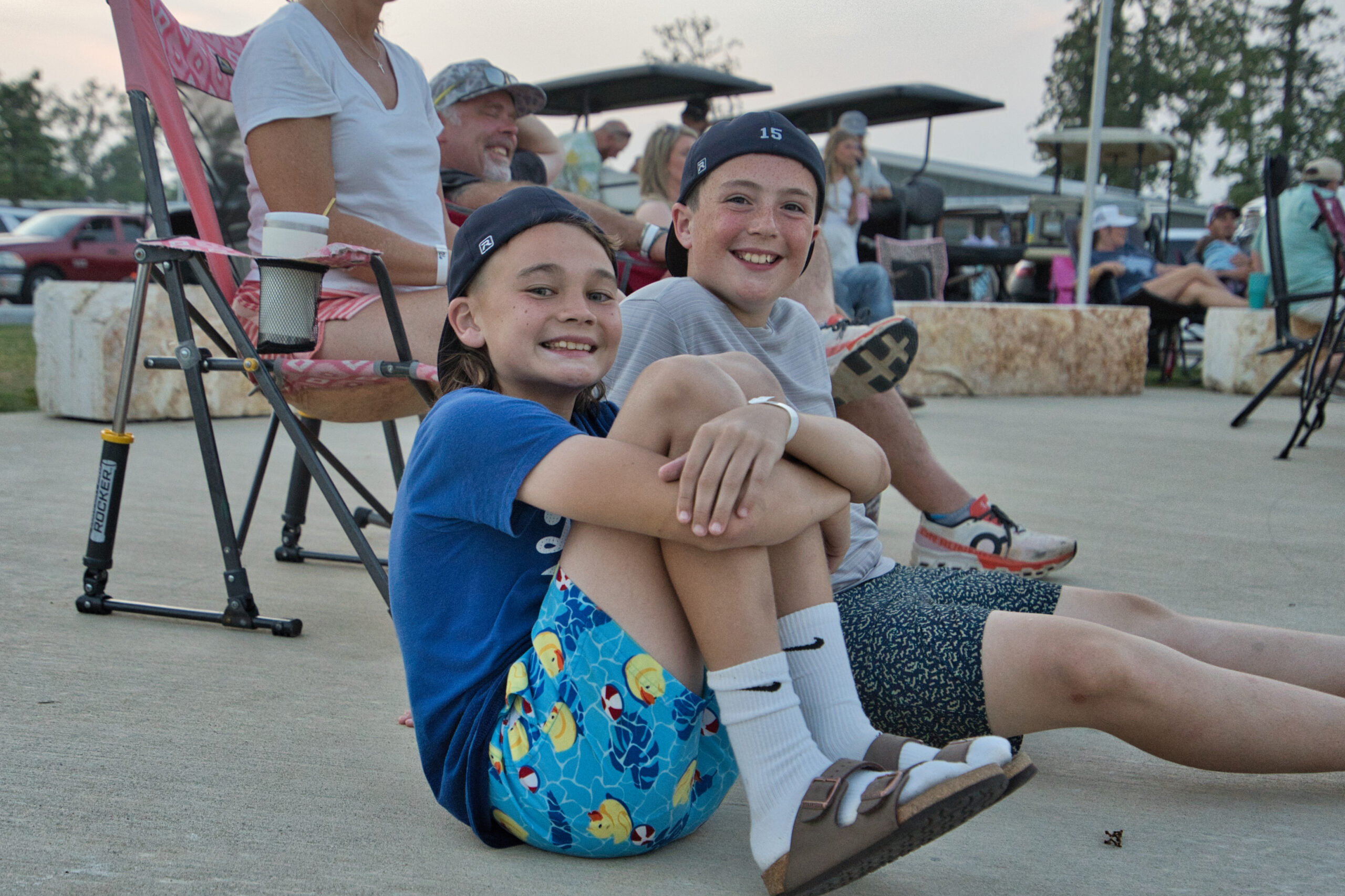 Two smiling kids in casual clothes sit on the ground near adults at an outdoor event, with chairs, cars, and trees in the background.