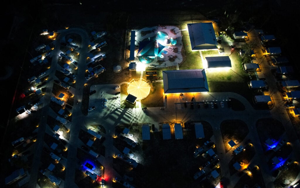 Aerial view of a lit-up campground at night with RVs, buildings, and a central illuminated area surrounded by parked vehicles.