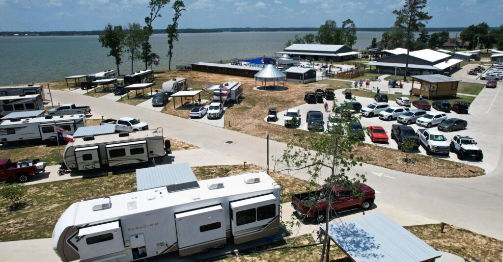 A lakeside RV park with several RVs, parked cars, a large gazebo, picnic areas, and buildings near the water under a partly cloudy sky.