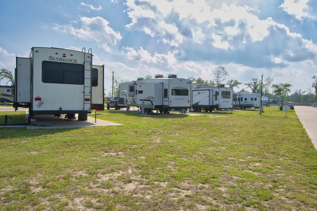 Several RVs are parked in designated spots on a grassy lot under a partly cloudy sky, with a paved road visible to the right.
